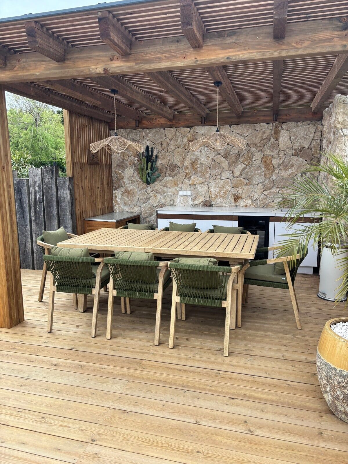 Table de jardin en bois et chaises vertes sous une pergola design avec mur en pierre et cuisine d'été extérieure.