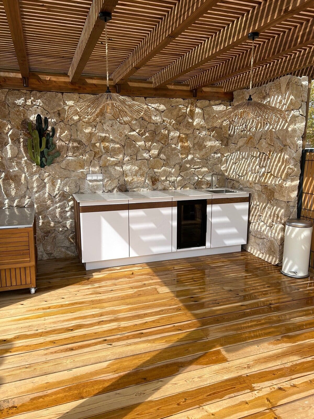 Cuisine d'été sous une pergola en bois avec mur en pierre rustique et mobilier moderne blanc.