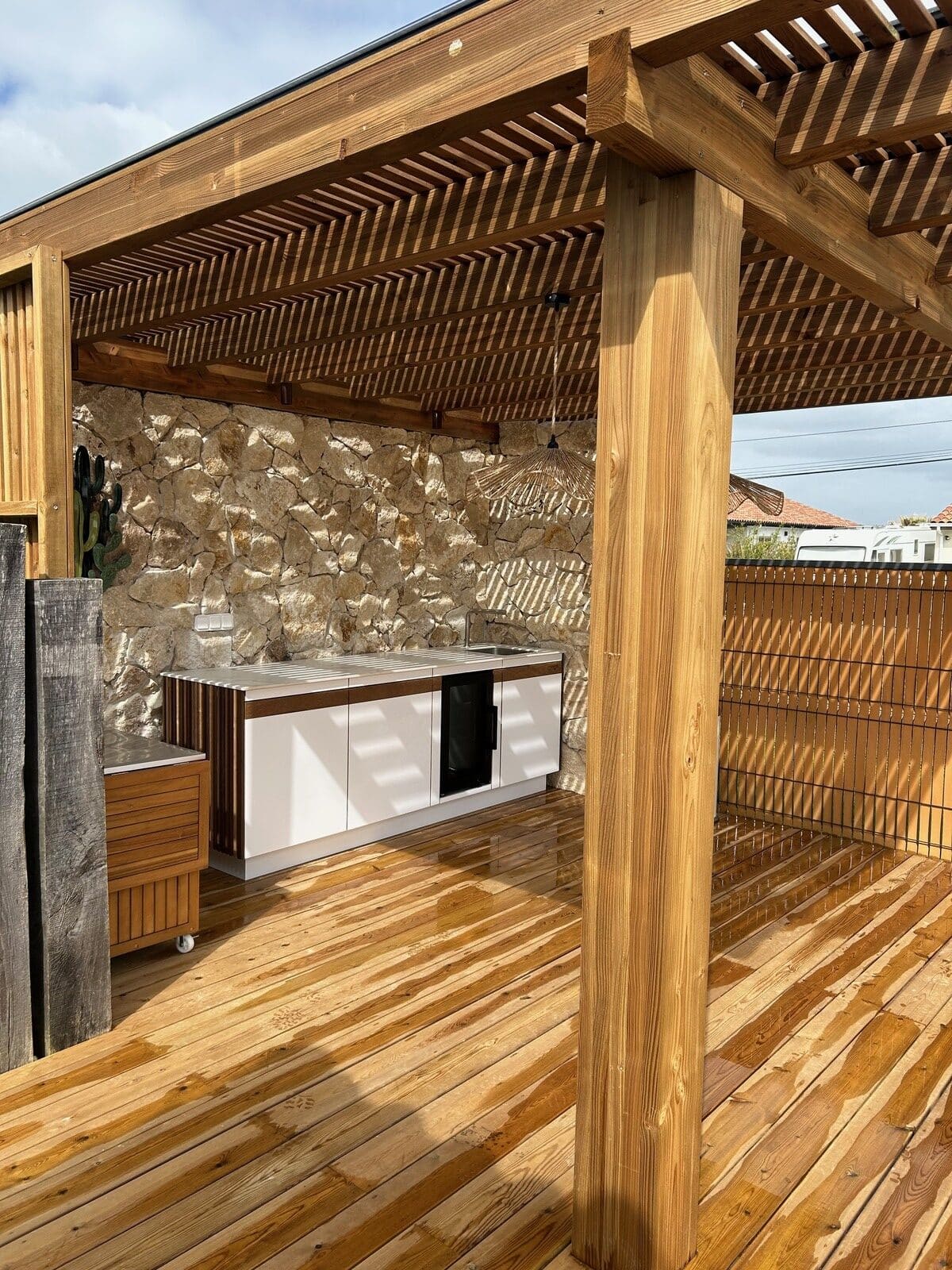 Cuisine extérieure moderne sous une pergola en bois avec mur en pierre naturelle et terrasse mouillée.