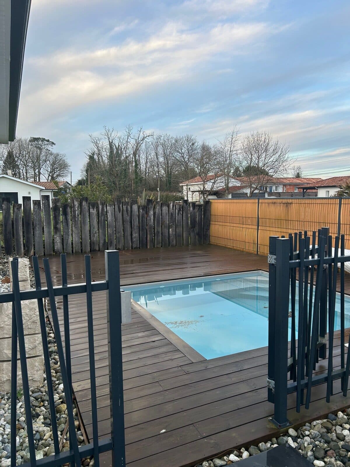 Piscine enterrée avec terrasse en bois sombre et clôture de sécurité moderne dans un jardin paysager.