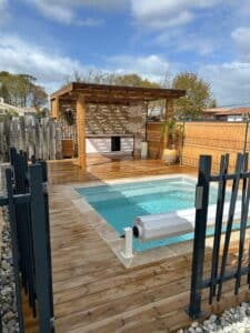 Piscine avec terrasse en bois, pergola design et cuisine extérieure devant un mur en pierre naturelle.