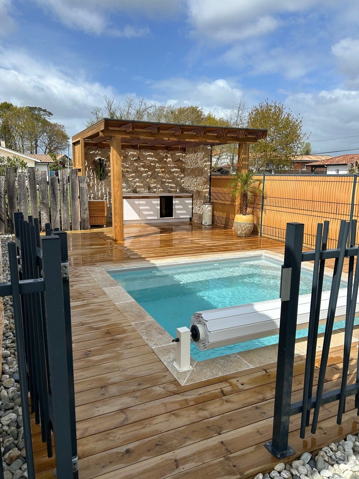 Piscine avec terrasse en bois, pergola design et cuisine extérieure devant un mur en pierre naturelle.